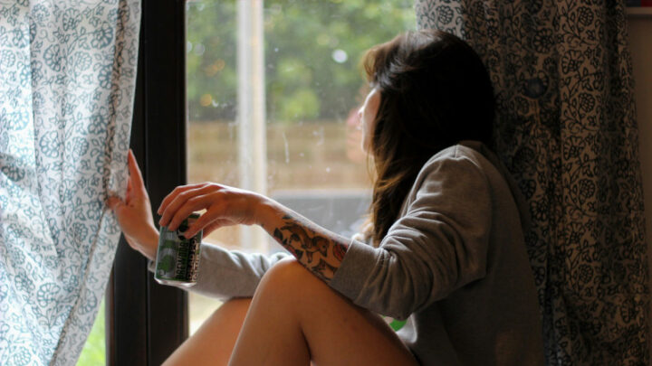 Kobieta uzależniona od alkoholu z puszką piwa w dłoni, zerka przez okno